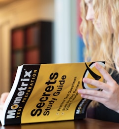 A person with long, wavy hair is holding a book titled 'Mometrix Test Preparation: Secrets Study Guide'. The cover is bright yellow with black text. The background appears blurred, focusing on the book.