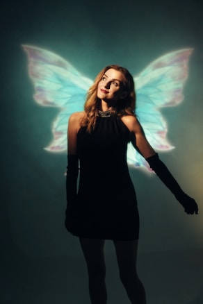 A woman stands gracefully in a dark setting wearing a black dress and long black gloves. Behind her, luminous wings with shades of blue and pink create the illusion of a fairy or angelic figure, adding a dreamy and magical element.