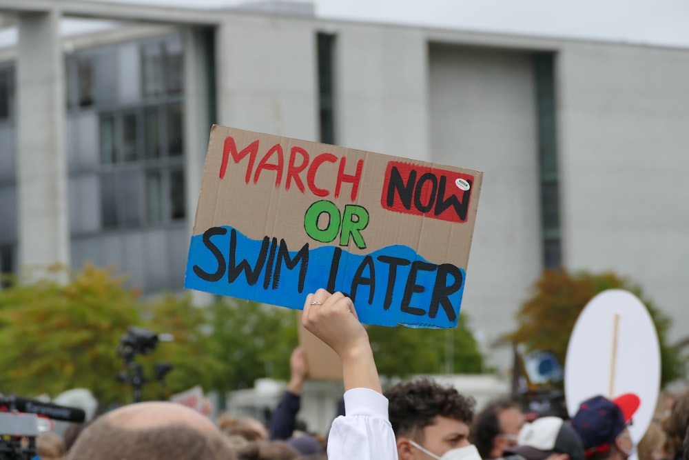 a person holding a sign that says march now or swim later
