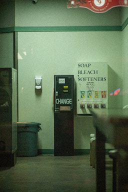 A dimly lit laundromat corner featuring a vending machine offering toiletries such as soap, bleach, and softeners. There is a change machine next to it, and a trash can is visible on the left side. The walls are plain with a hand sanitizer dispenser mounted on them.