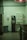 A dimly lit laundromat corner featuring a vending machine offering toiletries such as soap, bleach, and softeners. There is a change machine next to it, and a trash can is visible on the left side. The walls are plain with a hand sanitizer dispenser mounted on them.
