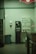 A dimly lit laundromat corner featuring a vending machine offering toiletries such as soap, bleach, and softeners. There is a change machine next to it, and a trash can is visible on the left side. The walls are plain with a hand sanitizer dispenser mounted on them.