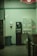 A dimly lit laundromat corner featuring a vending machine offering toiletries such as soap, bleach, and softeners. There is a change machine next to it, and a trash can is visible on the left side. The walls are plain with a hand sanitizer dispenser mounted on them.