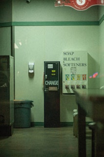 A dimly lit laundromat corner featuring a vending machine offering toiletries such as soap, bleach, and softeners. There is a change machine next to it, and a trash can is visible on the left side. The walls are plain with a hand sanitizer dispenser mounted on them.