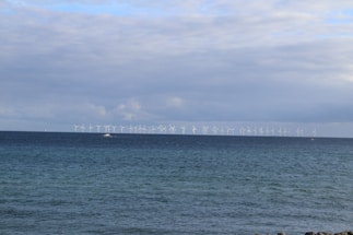 A calm seascape with numerous wind turbines positioned along the horizon. The waters are a deep blue, and the sky is partially covered with clouds, suggesting an overcast day. A lone boat is visible in the water, adding a sense of scale to the scene.