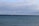 A calm seascape with numerous wind turbines positioned along the horizon. The waters are a deep blue, and the sky is partially covered with clouds, suggesting an overcast day. A lone boat is visible in the water, adding a sense of scale to the scene.