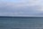 A calm seascape with numerous wind turbines positioned along the horizon. The waters are a deep blue, and the sky is partially covered with clouds, suggesting an overcast day. A lone boat is visible in the water, adding a sense of scale to the scene.
