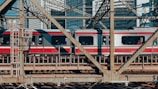 A red and silver passenger train travels over a steel bridge, with urban skyscrapers visible in the background. The geometric patterns of the bridge's metal beams frame the train as it moves through a bustling city environment.