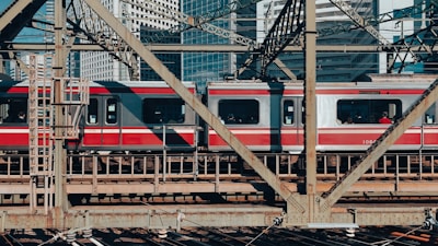 A red and silver passenger train travels over a steel bridge, with urban skyscrapers visible in the background. The geometric patterns of the bridge's metal beams frame the train as it moves through a bustling city environment.