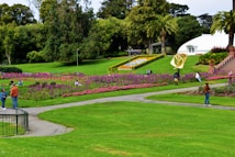 A landscaped park with vibrant flower beds featuring pink and purple blooms. People are strolling and relaxing on the grass. There are palm trees and a stairway on the right leading to a white domed building in the background. The scene is lush and well-maintained, conveying a peaceful public space.