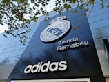 A modern building facade featuring a large emblem and the words 'Tienda Bernabéu'. The prominent logo is above the shop name, and the brand 'Adidas' is displayed below. Trees with green leaves are visible in the top part of the image.