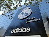 A modern building facade featuring a large emblem and the words 'Tienda Bernabéu'. The prominent logo is above the shop name, and the brand 'Adidas' is displayed below. Trees with green leaves are visible in the top part of the image.