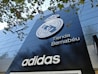 A modern building facade featuring a large emblem and the words 'Tienda Bernab&eacute;u'. The prominent logo is above the shop name, and the brand 'Adidas' is displayed below. Trees with green leaves are visible in the top part of the image.