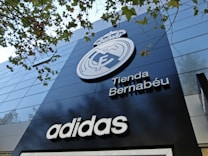 A modern building facade featuring a large emblem and the words 'Tienda Bernab&eacute;u'. The prominent logo is above the shop name, and the brand 'Adidas' is displayed below. Trees with green leaves are visible in the top part of the image.