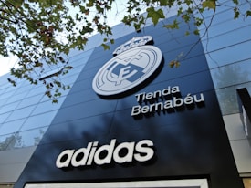 A modern building facade featuring a large emblem and the words 'Tienda Bernabéu'. The prominent logo is above the shop name, and the brand 'Adidas' is displayed below. Trees with green leaves are visible in the top part of the image.