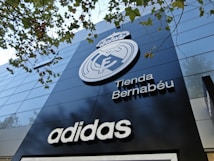 A modern building facade featuring a large emblem and the words 'Tienda Bernab&eacute;u'. The prominent logo is above the shop name, and the brand 'Adidas' is displayed below. Trees with green leaves are visible in the top part of the image.