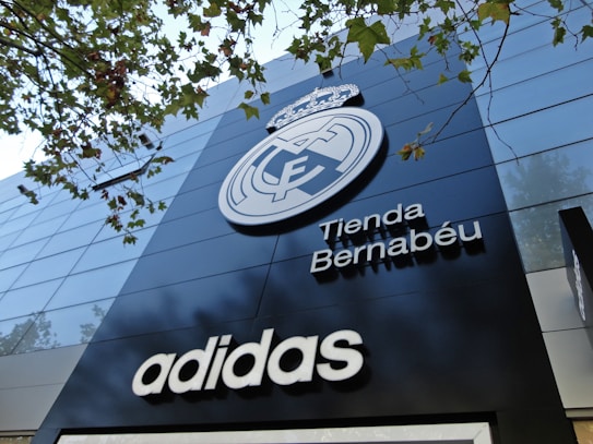 A modern building facade featuring a large emblem and the words 'Tienda Bernab&eacute;u'. The prominent logo is above the shop name, and the brand 'Adidas' is displayed below. Trees with green leaves are visible in the top part of the image.