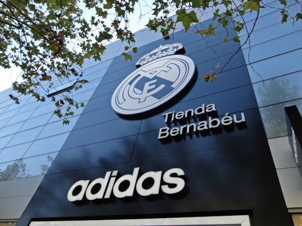 A modern building facade featuring a large emblem and the words 'Tienda Bernab&eacute;u'. The prominent logo is above the shop name, and the brand 'Adidas' is displayed below. Trees with green leaves are visible in the top part of the image.