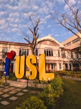 A large sign with the letters 'USL' stands prominently in a landscaped area with green grass and plants. The building behind the sign is an older architectural style with white and red details. The sky is blue with scattered clouds, and there is a leafless tree in the foreground.