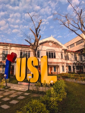 A large sign with the letters 'USL' stands prominently in a landscaped area with green grass and plants. The building behind the sign is an older architectural style with white and red details. The sky is blue with scattered clouds, and there is a leafless tree in the foreground.