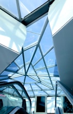 A geometric glass ceiling with a steel frame, showcasing a modern architectural design. Below, escalators are visible, suggesting a commercial or public space. The overhead glass panels allow natural light to illuminate the area.