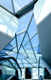 A geometric glass ceiling with a steel frame, showcasing a modern architectural design. Below, escalators are visible, suggesting a commercial or public space. The overhead glass panels allow natural light to illuminate the area.