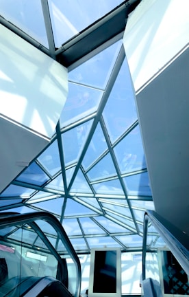 A geometric glass ceiling with a steel frame, showcasing a modern architectural design. Below, escalators are visible, suggesting a commercial or public space. The overhead glass panels allow natural light to illuminate the area.