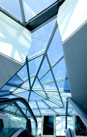 A geometric glass ceiling with a steel frame, showcasing a modern architectural design. Below, escalators are visible, suggesting a commercial or public space. The overhead glass panels allow natural light to illuminate the area.