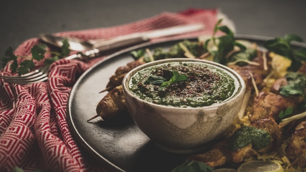 A bowl of green chutney garnished with mint on a dark plate, surrounded by skewered grilled meat pieces and fresh herbs. Yellow rice and a patterned red cloth with cutlery are also visible, creating a rustic and appetizing setting.