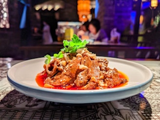 A white plate holding a savory dish garnished with coriander is placed on a table. The dish consists of tender, saucy pieces of meat topped with sesame seeds and nuts. The background is slightly out of focus, revealing a dimly lit dining environment with warm lighting and people seated at a table.