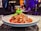 A white plate holding a savory dish garnished with coriander is placed on a table. The dish consists of tender, saucy pieces of meat topped with sesame seeds and nuts. The background is slightly out of focus, revealing a dimly lit dining environment with warm lighting and people seated at a table.