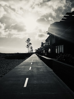 A long, straight path or road leading towards the horizon under a bright sun with rays streaming down. On the right side are silhouettes of modern buildings, and on the left, palm trees stand tall against the sky. The scene is predominantly in monochrome, emphasizing shadows and light.