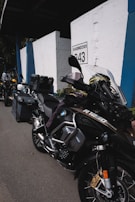 A line of motorcycles is parked along a street near a wall with a sign displaying '04BND201 P43'. The motorcycles are equipped with large side cases and one sports a significant R1250 marking. The scene is under a covered area with the wall painted blue and white.