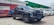 A modern black SUV is parked on a paved area in front of a red advertisement banner. Nearby, there are other cars in white and blue. The setting appears to be an open-air dealership or showroom with a partially visible green canopy in the background.