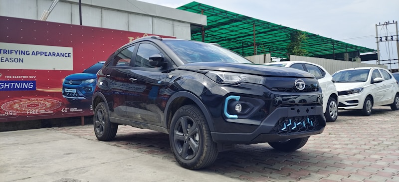 A modern black SUV is parked on a paved area in front of a red advertisement banner. Nearby, there are other cars in white and blue. The setting appears to be an open-air dealership or showroom with a partially visible green canopy in the background.