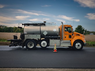 A bright orange roadwork truck with a large metal tank is parked on a freshly paved road. The vehicle has two hazard cones placed near its front wheel. In the background, a rural landscape with fields and a line of trees is visible under a partly cloudy sky.