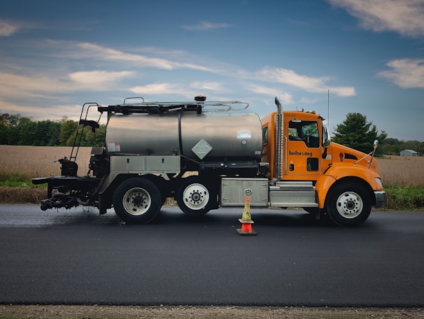 A bright orange roadwork truck with a large metal tank is parked on a freshly paved road. The vehicle has two hazard cones placed near its front wheel. In the background, a rural landscape with fields and a line of trees is visible under a partly cloudy sky.