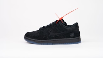 A black sneaker with a sleek design featuring a red tag attached near the laces. It has a perforated texture and is positioned against a plain white background. A subtle logo with five strike-through lines is visible on the side.