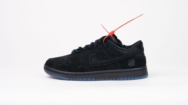 A black sneaker with a sleek design featuring a red tag attached near the laces. It has a perforated texture and is positioned against a plain white background. A subtle logo with five strike-through lines is visible on the side.