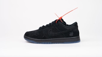 A black sneaker with a sleek design featuring a red tag attached near the laces. It has a perforated texture and is positioned against a plain white background. A subtle logo with five strike-through lines is visible on the side.