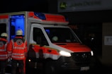 An ambulance with the Deutsches Rotes Kreuz insignia is parked with its rear door open, revealing a dimly lit blue interior. Two individuals in reflective emergency uniforms and helmets stand near the back of the vehicle.