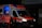 An ambulance with the Deutsches Rotes Kreuz insignia is parked with its rear door open, revealing a dimly lit blue interior. Two individuals in reflective emergency uniforms and helmets stand near the back of the vehicle.