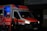 An ambulance with the Deutsches Rotes Kreuz insignia is parked with its rear door open, revealing a dimly lit blue interior. Two individuals in reflective emergency uniforms and helmets stand near the back of the vehicle.