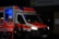 An ambulance with the Deutsches Rotes Kreuz insignia is parked with its rear door open, revealing a dimly lit blue interior. Two individuals in reflective emergency uniforms and helmets stand near the back of the vehicle.