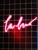 A glowing neon pink sign with a cursive script hangs against a black tiled wall with thin orange lines. The section of the wall illuminated by the sign showcases a warm and vibrant glow.