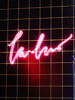 A glowing neon pink sign with a cursive script hangs against a black tiled wall with thin orange lines. The section of the wall illuminated by the sign showcases a warm and vibrant glow.