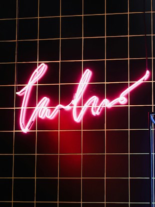 A glowing neon pink sign with a cursive script hangs against a black tiled wall with thin orange lines. The section of the wall illuminated by the sign showcases a warm and vibrant glow.