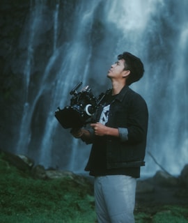 A person stands outdoors holding a professional camera against a backdrop of a cascading waterfall. The individual appears focused and is dressed casually in a denim jacket and light-colored pants. The lush greenery at the base of the waterfall adds to the serene natural setting.