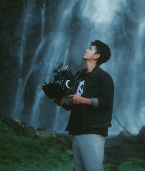 A person stands outdoors holding a professional camera against a backdrop of a cascading waterfall. The individual appears focused and is dressed casually in a denim jacket and light-colored pants. The lush greenery at the base of the waterfall adds to the serene natural setting.