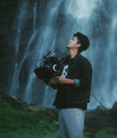 A person stands outdoors holding a professional camera against a backdrop of a cascading waterfall. The individual appears focused and is dressed casually in a denim jacket and light-colored pants. The lush greenery at the base of the waterfall adds to the serene natural setting.